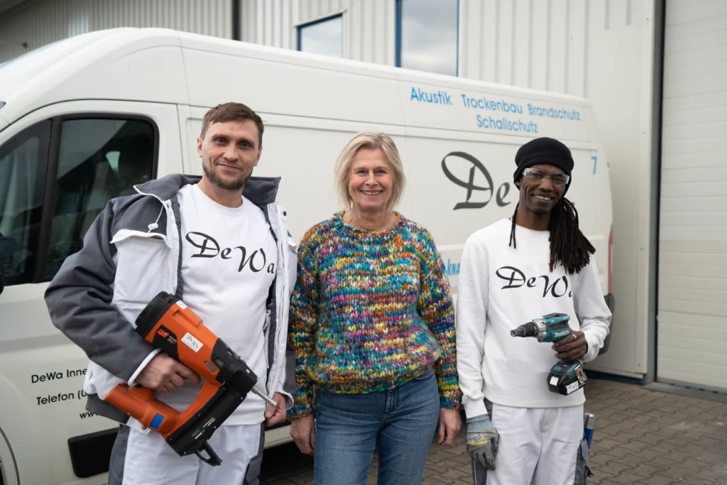Zwei Handwerker von DeWa Innenausbau mit einer Frau vor einem Firmenfahrzeug, ausgestattet mit Werkzeug und Arbeitskleidung, bereit für den nächsten Einsatz.